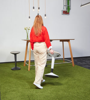 Woman carrying white Younit sit stand stool to standing desk.